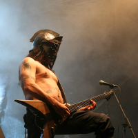 A shirtless musician wearing a metallic helmet with horn-like protrusions plays an electric guitar on stage at the 2011 Kilkim Žaibu festival. The stage is filled with a smoky haze, and microphones are positioned in front of him, while bright stage lights and equipment form the backdrop.
