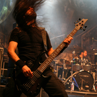 A musician with long hair headbangs while playing a black electric guitar on stage at Kilkim Žaibu festival. He is dressed in black and has a wristband on his left arm. In the background, a drummer is visible with a drum kit. Stage lights and a slightly smoky atmosphere contribute to the intense concert setting.