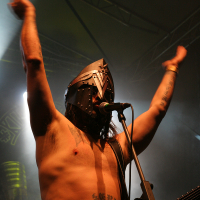 A shirtless musician with tattoos is performing on stage at Kilkim Žaibu 2011. He is wearing a black metal helmet with intricate designs. His arms are raised in the air as he sings into a microphone mounted on a stand. Dark stage lighting and smoke create a dramatic atmosphere around him. A guitar is strapped over his shoulder.