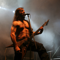 A shirtless man with long hair and tattoos is playing a V-shaped electric guitar on stage at the 2011 Kilkim Žaibu festival. He is singing into a microphone, surrounded by dramatic lighting and smoke effects. Wearing dark pants, the stage is equipped with amplifiers, one of which is branded "Marshall.