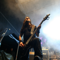 A musician with long hair performs energetically on a fog-filled stage at Kilkim Žaibu 2011, playing an electric guitar. He stands in front of a stack of amplifiers, with bright lights and equipment surrounding him. The scene captures the raw energy of a live rock concert.