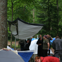 A group of people are camping in a forested area, surrounded by trees and tents at the 2011 Kilkim Žaibu festival. One person is seen hanging a tent in an unusual upside-down position from a tree. Several others are standing and conversing nearby, while someone in a red jacket crouches close to a tent in the foreground.