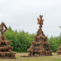 Three large, pyramid-shaped bonfires made from logs and covered in straw stand in a grassy field. Each bonfire, reminiscent of the 2012 Kilkim Žaibu festival, has a unique decoration at the top: one features a diamond shape, another has a human-like figure, and the third has a simple peak. Trees and shrubs are visible in the background under a cloudy sky.
