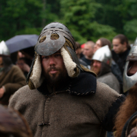 A bearded man dressed in medieval attire and wearing a metal helmet with fur lining stands among a group of similarly dressed individuals. He looks directly at the camera. The background is filled with several people, some wearing helmets, suggesting a historical reenactment event at the 2012 Kilkim Žaibu festival, set outdoors.