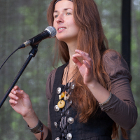 A woman with long, reddish-brown hair wears a black dress adorned with multiple buttons and a brown belt. She stands in front of a microphone on a stand, her hand raised slightly as if gesturing while speaking or singing. The blurred foliage suggests an outdoor setting, reminiscent of Kilkim Žaibu 2012.