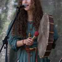 A woman with long curly hair, wearing a green dress with wooden buttons, performs on stage at Kilkim Žaibu 2012. She is holding a large, round hand drum and a red-tipped drumstick. Standing in front of a microphone, she smiles as she sings or speaks. The background is speckled with light.