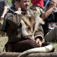 A man sits on the ground wearing medieval-style clothing with a fur draped over his shoulders. He holds one hand to his face and has a thoughtful expression. Surrounded by others, some clapping, the scene appears to be from the 2012 Kilkim Žaibu festival, set in a grassy reenactment area outdoors.