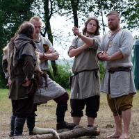 Four men in historical attire stand outdoors at a festival, engaged in conversation. They wear tunics, belts, and trousers typical of medieval or Viking reenactments. One man gestures while others listen attentively. The backdrop featuring trees, grass, and a portable toilet suggests a rural or event setting like Kilkim Žaibu 2012.