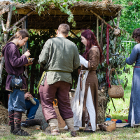 A group of five people in medieval attire stands and sits by a rustic shelter made of branches and straw in a lush, green outdoor setting at the 2012 Kilkim Žaibu festival. They appear to be engaged in various activities, with some looking down at objects in their hands or sitting inside the shelter.