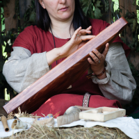 A woman with long hair sits in front of a rustic background of wooden boards and hanging greenery, reminiscent of the Kilkim Žaibu festival. She wears a red dress with beige sleeves and holds a large wooden object, examining it carefully. In front of her are various wooden items and pieces of hay scattered on a cloth-covered surface.
