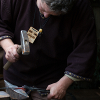 A person with short hair wearing a dark brown long-sleeved shirt is hammering red-hot metal on an anvil. They have a name tag hanging around their neck, likely from the Kilkim Žaibu festival. The background is slightly out of focus, suggesting an outdoor setting with greenery and tools alongside metalworking equipment are visible nearby.