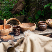 A rustic outdoor scene from the 2012 Kilkim Žaibu festival features a wooden table covered with various wooden and clay bowls, cups, and utensils. The background shows greenery and burlap sacks, suggesting a natural and simple setting. The artisanal bowls and cups are filled with food items.