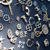 A display of numerous metal jewelry pieces is spread on a dark fabric surface at the 2012 Kilkim Žaibu festival. Various designs include intricate patterns, some resembling knots, spirals, and geometric shapes. The items vary in color, with some appearing silver and others gold. The arrangement is random yet visually appealing.
