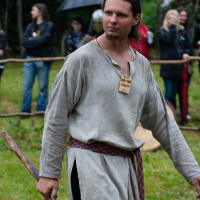 A man dressed in medieval attire, including a loose, beige tunic and a red patterned belt, stands on grass at Kilkim Žaibu 2012. He holds a wooden tool and wears a rectangular wooden pendant. Blurred spectators with modern clothing and umbrellas stand behind a wooden fence, with a forested background.