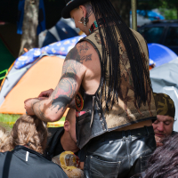 A person with long dreadlocks, a top hat, and detailed tattoos on their arms leans over a group seated at a table outdoors. Sporting a sleeveless vest with a Native American motif and black leather pants, they evoke the wild spirit of Kilkim Žaibu 2012. Tents and trees in the background suggest a campsite setting.