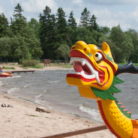 A brightly painted dragon boat with a yellow, red, and green dragon head rests on a sandy beach by a lake during the lively 2012 Kilkim Žaibu festival. Several other colorful boats are in the background near the shoreline. Tall trees and a partly cloudy sky are visible in the distance, completing this vivid scene.