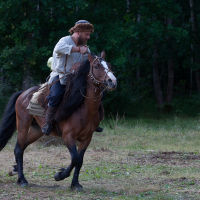 A man in medieval attire rides a brown horse through a grassy field at the 2012 Kilkim Žaibu festival. He is wearing a white shirt, blue pants, and a fur cap. The horse has a white blaze on its face and a brown saddle blanket. Trees form a dense background at the edge of the field, seemingly in motion, heading left.