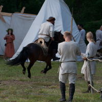 A scene of medieval reenactment at Kilkim Žaibu 2012 shows people dressed in period costumes. In the foreground, two men in beige tunics and boots stand, one holding a spear. A man on horseback rides past them. In the background, a woman in a long dress stands near a white canvas tent. Green trees surround the area.