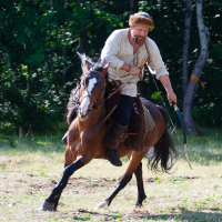 A bearded man wearing rustic clothing rides a brown horse at full gallop through a wooded area, gripping a sword in his right hand. The horse appears alert and energetic. In the background, dense trees and a stack of hay evoke an adventurous, historical setting reminiscent of the 2012 Kilkim Žaibu festival.