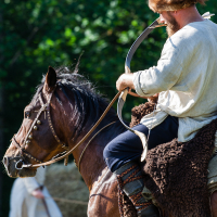 A man dressed in medieval attire, including a fur hat and a light tunic, is riding a brown horse. He holds a curved bow, ready to shoot. The scene, reminiscent of Kilkim Žaibu 2012, unfolds in a lush, green forest with another person in similar attire visible in the distance, partially obscured.