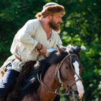 A man dressed in medieval attire with a fur hat and white tunic rides a brown horse with a white stripe on its face. He looks focused and determined, holding the reins with both hands. The background, reminiscent of Kilkim Žaibu 2012, is a lush forest with dense green foliage, perfect for the festival vibe.