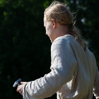 A person with long, light hair tied back, wearing a light-colored tunic, stands outdoors with trees in the background at Kilkim Žaibu 2012. The individual holds a sword in their right hand, the blade pointed downwards. The scene is set in daylight, highlighting the person's back and right side.