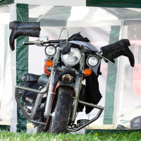 A motorcycle is parked on grassy ground inside a white and green canopy tent at the Kilkim Žaibu festival. The bike is partially covered with a black jacket draped over the handlebars, and black motorcycle chaps hanging from either side of the tent frame. The background shows green foliage and a red vehicle.
