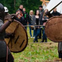 Two people clad in medieval armor and carrying large round shields engage in a mock sword fight at the Kilkim Žaibu festival. The audience, dressed in modern casual attire, watches intently. The scene takes place outdoors with greenery and a rustic tent visible in the background, evoking the spirit of 2012.