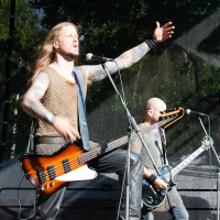 Two men perform on a stage at Kilkim Žaibu 2012, both wearing chainmail armor. The lead performer, with long hair and tattoos, energetically sings into a microphone with his left arm raised. He also plays a bass guitar. The other man, bald, plays an electric guitar in the background. Trees are visible outdoors.