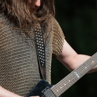 At Kilkim Žaibu 2012, a person with long hair is playing an electric guitar outdoors. They're sporting a chainmail shirt and a studded leather strap for the guitar, with a festival wristband visible on their wrist. The dark, blurred background draws attention to them and the instrument.