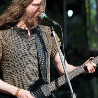 A musician with long hair performs on stage at the Kilkim Žaibu festival, wearing a metal chainmail tunic. He plays a black electric guitar and sings into a microphone. The background shows stage lighting and equipment, lit by natural daylight, suggesting an outdoor event in 2012.