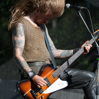 A long-haired musician, wearing a sleeveless chainmail shirt and black leather pants, energetically plays an electric bass guitar on stage at Kilkim Žaibu 2012. His head is down, hair flying mid-air in a dynamic scene. Tattoos cover his arms, and a microphone stand is positioned to his right.