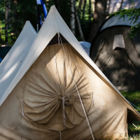 A beige canvas tent is set up on grassy terrain in a wooded area. The tent is closed at the front, with ropes securing the entrance. Another similar tent and trees are visible in the background, under sunlight filtering through the leaves. The scene suggests a campground reminiscent of Kilkim Žaibu 2012.