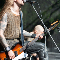 At the 2012 Kilkim Žaibu festival, a musician with long blonde hair and tattoos passionately sings into a microphone while playing a brown electric guitar on stage. He wears a chainmail shirt and black wristbands. Another band member with a shaved head is seen playing an instrument in the background.