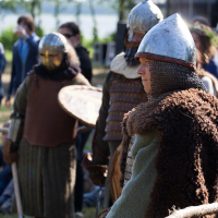 People dressed in medieval warrior costumes, featuring chainmail, helmets, and fur, stand outdoors among trees and grass. Some carry shields, while one focuses attentively to their right. The background is slightly blurred, showing other individuals and the natural setting of the 2012 Kilkim Žaibu festival.