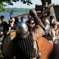 At a 2012 festival, two medieval re-enactors wearing chainmail and helmets engage in combat with shields and axes at an outdoor event. Onlookers in modern clothing surround them, watching intently. The backdrop includes a large tree, a tent, and the serene body of water visible in the distance.