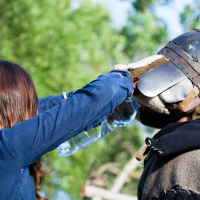 Attending the Kilkim Žaibu festival in 2012, a person with long brown hair, wearing a blue jacket, pours water from a plastic bottle over the mouthguard of a medieval helmet worn by another individual in armor. The background is blurred greenery, suggesting an outdoor setting.