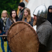 A bearded individual dressed in medieval armor, including a chainmail suit and a shiny helmet, holds a large leather shield and sword. They stand outdoors at the 2012 Kilkim Žaibu festival with a backdrop of blurred onlookers and a white tent, capturing the essence of this historical reenactment event.