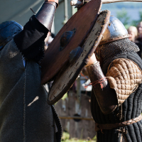 Two individuals engage in a medieval combat reenactment outdoors at Kilkim Žaibu 2012. Both wear period-appropriate clothing and metal helmets. They clash with one another; one raises a sword, and the other defends with a large round shield. Onlookers in the background watch the intense mock battle.