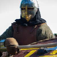 A person dressed in medieval armor, including a metal helmet with a visor, holds a shield adorned with yellow markings on a red background. Wearing a brown leather chest piece over a dark hood and green garment, the individual stands against a blurred outdoor background at the 2012 Kilkim Žaibu festival.