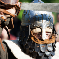 A person wearing medieval armor, including a scaled helmet with face protection and a padded collar, holds up a sword at Kilkim Žaibu 2012. The intricate helmet features metal plates and leather accents. The armor, detailed and well-crafted, gleams in the sunlight with a blurred outdoor background.