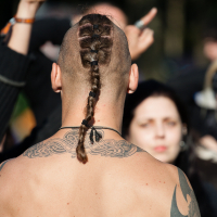 A shirtless person with intricate back tattoos and a long braided ponytail stands with their back to the camera at Kilkim Žaibu 2012. They are outdoors among a crowd, where some people are raising their hands. The background is slightly blurred, focusing on the person in the foreground and highlighting their hairstyle and tattoos.