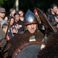 A person dressed as a medieval warrior, wearing a helmet and chainmail, holds a sword in one hand and a shield in the other. They appear to be in mid-battle during the 2012 Kilkim Žaibu reenactment event. The background shows a crowd of onlookers, many in casual clothing, observing the action.