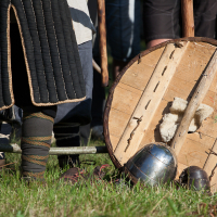 A medieval reenactor stands in a grassy field at Kilkim Žaibu 2012, with quilted leg armor and wrapped boots. At their feet are a wooden round shield, a metal helmet, and two small metal objects. The scene captures elements of historical combat attire and equipment.