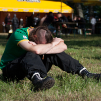 A person in a green shirt and black pants sits on the grass with their head resting on crossed arms and knees drawn up. The background features a yellow tent with "2012 Kilkim Žaibu" branded on it, and several people standing near it. The setting appears to be an outdoor event or festival.