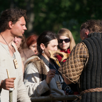 A group of people dressed in medieval clothing participate in the 2012 Kilkim Žaibu outdoor event. One man on the left holds a staff, while a woman in the center looks at a piece of parchment. Another person facing away from the camera wears a quilted garment with a rope belt. Other participants and onlookers are around.