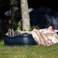 Two people are lying down on an inflatable mattress, covered with a blanket featuring leaf patterns. The mattress is placed outdoors on the grass, beneath trees with two tents visible behind them. The scene, reminiscent of Kilkim Žaibu 2012, captures the essence of a peaceful camping setting in daylight.