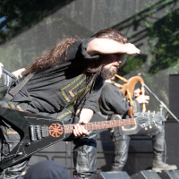 A musician with a long beard and long hair, wearing a black outfit, plays an electric guitar on an outdoor stage. He is shielding his eyes from the sun with his hand. Behind him, another musician plays the bagpipes. The audience at Kilkim Žaibu 2012 appears to be watching the performance intently.
