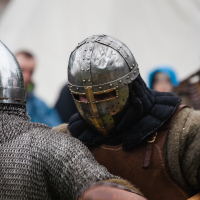 At the 2012 Kilkim Žaibu festival, two medieval reenactors donning chainmail and metal helmets engage in a close-combat fight. The fighter in the foreground wields a weapon, while the other, partially turned away, wears a helmet with a cross-shaped visor. A tent and blurred spectators are visible in the background.