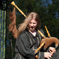 A musician with long blond hair plays the bagpipes on an outdoor stage at Kilkim Žaibu 2012. They wear a black, traditionally styled tunic with decorative patterns. The bagpipes feature a wooden, natural finish and a decorative tassel. Trees and stage lighting are visible in the background.